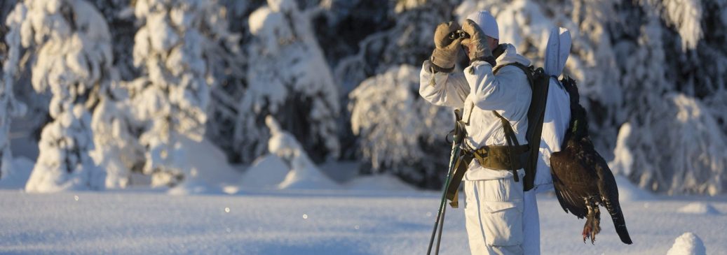 Lumipukuinen metsästäjä kiikaroi.