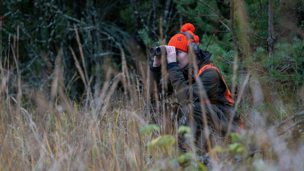Oranssipipoinen metsästävä nainen kiikaroi metsän reunasta kuulosuojaimet korvillaan ja radiopuhelin taskussaan.
