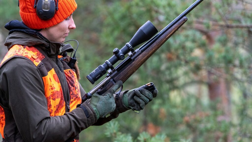 Oranssiliivinen metsästävä nainen laittaa patruunoilla ladattua lipasta kiinni kivääriin kuulosuojaimet korvillaan.
