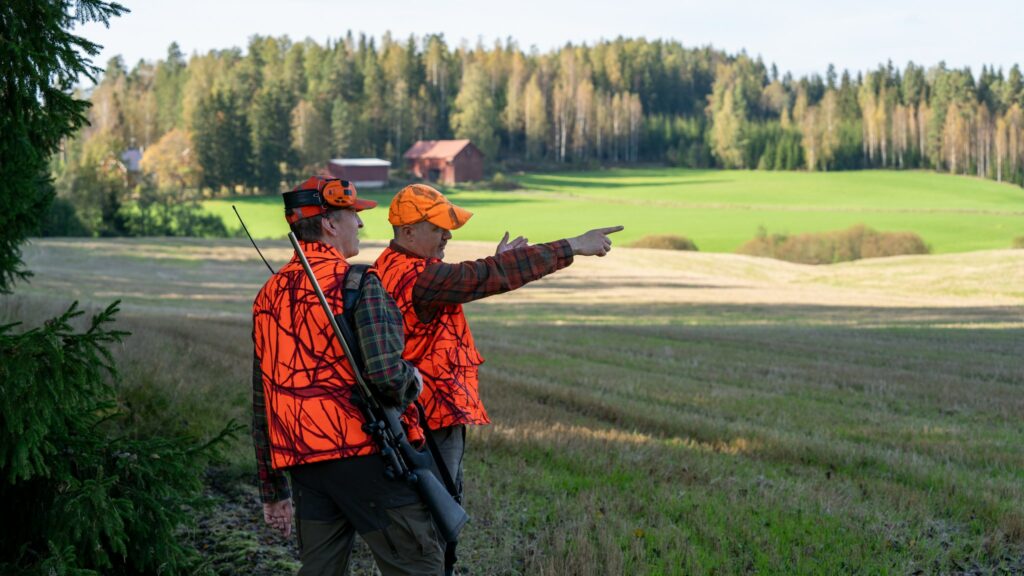 Metsästyksenjohtaja osoittaa ampumasektorin toiselle metsästäjälle. Taustalla talo.