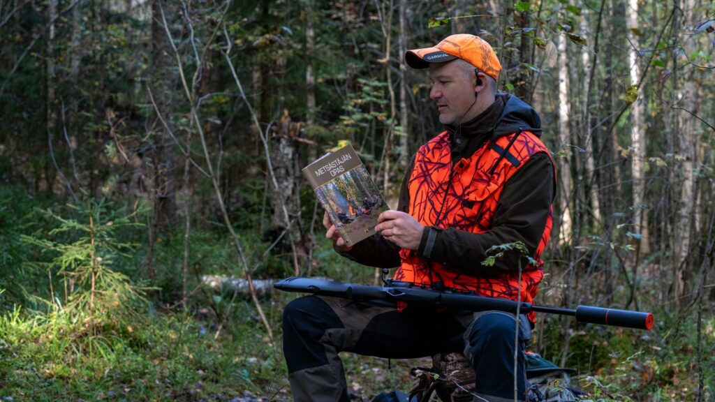 En jägare klädd i orangefärgad klädsel läser handboken med vapnet i famnen. 
