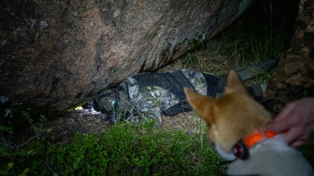 Metsästäjä lopettaa supikoiran kiven alle. Koira kytkettynä taustalla.